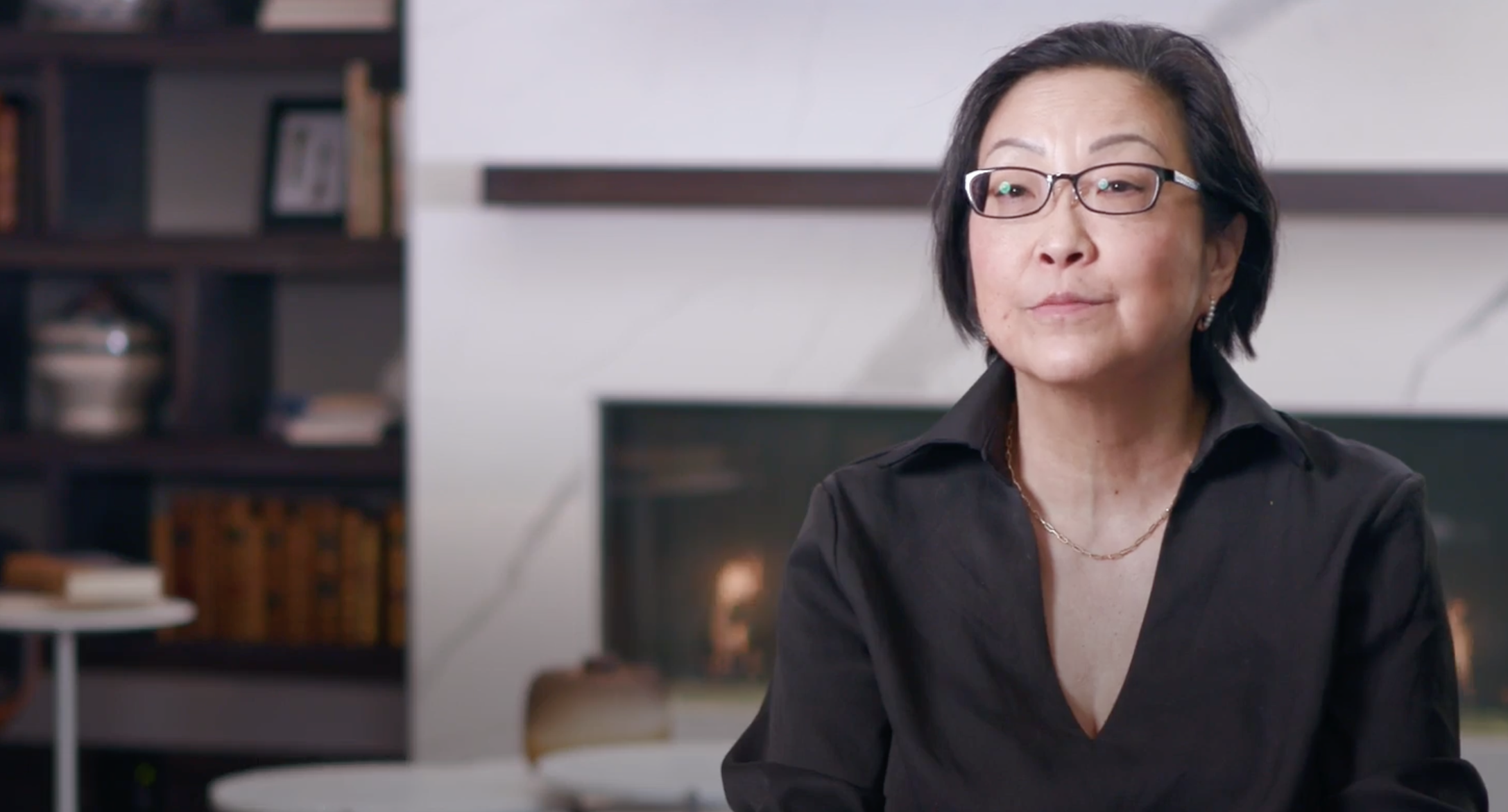 Whistleblower Aid CEO Libby Liu wears glasses and a black shirt with a simple gold chain. She is sitting in the forefront with a white marble fireplace and small, decorated book case in the background.
